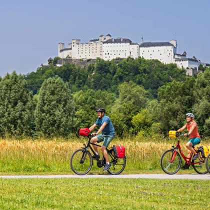 Drei Radfahrer mit gelben Taschen fahren auf einem Weg durch grüne Wiesen mit der Festung Hohensalzburg auf einem bewaldeten Hügel im Hintergrund.