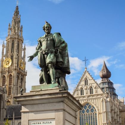 Bronzestatue von Peter Paul Rubens auf einem Sockel vor der Liebfrauenkathedrale in Antwerpen mit ihrem hohen gotischen Turm unter blauem Himmel.