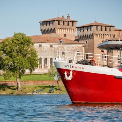 Rot-weißes Passagierschiff Ave Maria auf blauem Wasser vor historischen Festungsmauern und Türmen in Mantua mit grünen Bäumen am Ufer.