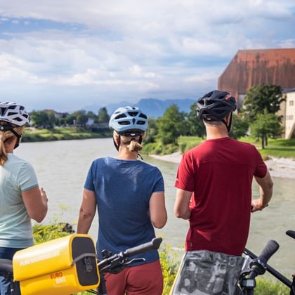 Three cyclists with helmets viewing the Salzach River near Laufen. A church with red roof and tower stands on the opposite riverbank.