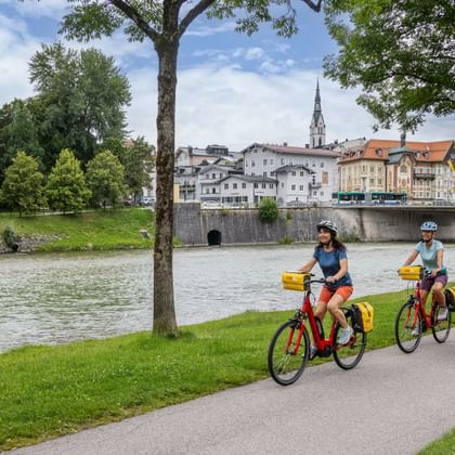 Three cyclists riding along the Isar River in Bad Tölz. Historic town buildings and church spire visible across the river under green trees.
