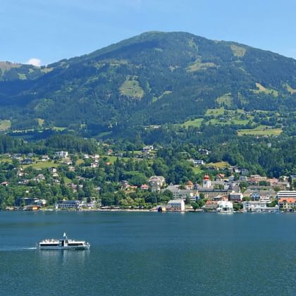 Passenger ferry crossing a Carinthian lake with a town on the shore and green mountains in the background under blue sky.
