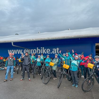 Large group of cyclists with bikes and yellow bags posing in front of blue Eurobike building during company trip along the Danube.