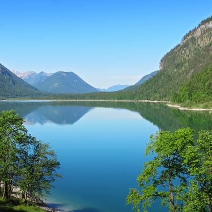Sylvensteinstausee umgeben von bewaldeten Bergen mit ruhigem blauen Wasser, das Himmel und Gipfel in den bayerischen Alpen spiegelt.