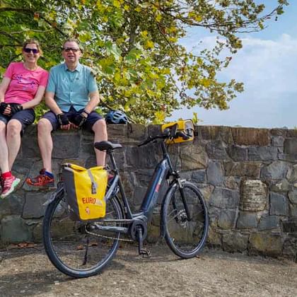 Zwei Radfahrer mit Sonnenbrillen sitzen auf einer Steinmauer mit ihrem geparkten Fahrrad davor, am Alpe Adria Radweg bei Grado.