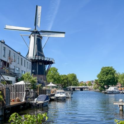 Traditionelle holländische Windmühle De Adriaan an einem Kanal in Haarlem mit Booten am Ufer und historischen Gebäuden unter blauem Himmel.
