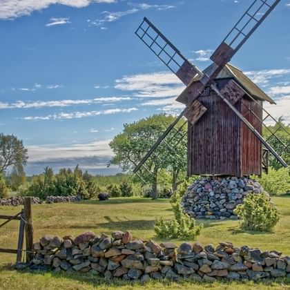 Wooden windmill on stone foundation in Latvia or Estonia, surrounded by stone walls, green meadows, and trees under blue sky.