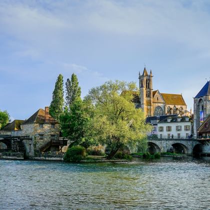 View of Moret-sur-Loing with stone bridge crossing the Loing river, historic church with tower, and traditional buildings under blue sky.