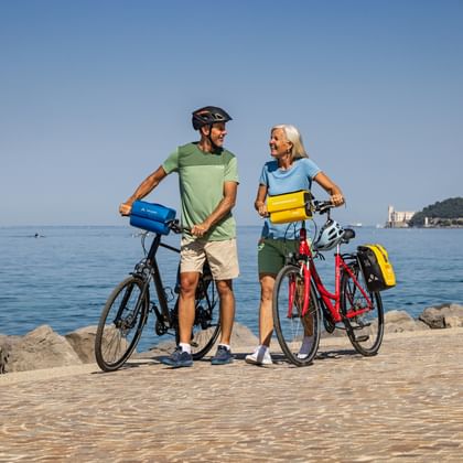 Paar mit Tourenrädern auf gepflastertem Uferweg bei Triest. Schloss Miramare auf entferntem Hügel über ruhiger blauer Adria sichtbar.