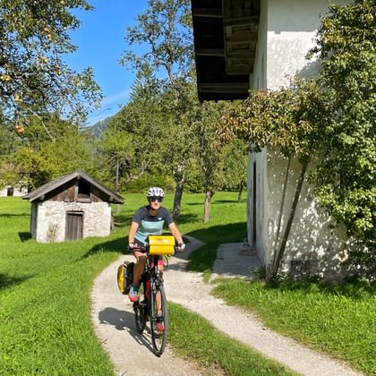 Cyclist with yellow panniers riding on a paved path through green meadows near Reit on the Tauern Radweg, with traditional buildings and trees.