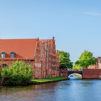 Rote Backstein-Salzspeicher am Kanal in Lübeck mit Kirchtürmen, Brücke und Ausflugsboot unter blauem Himmel.