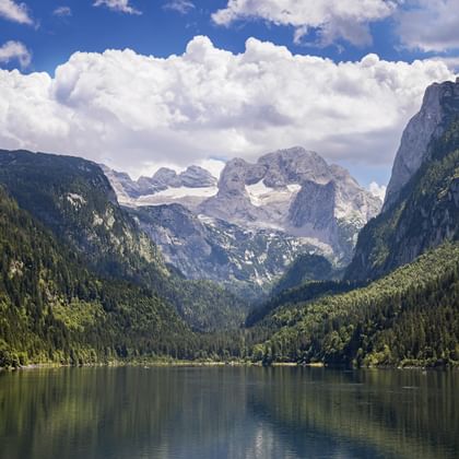 Lake Gosausee in Salzkammergut with forested mountains and Dachstein massif with snow-capped peaks under dramatic clouds.