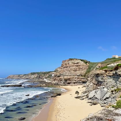 Sandstrand in der Bucht São Pedro da Cadeira mit geschichteten Felsklippen, Wellen am Ufer und Gebäuden auf dem Hügel unter blauem Himmel.