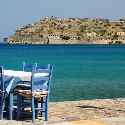 Blick über einen blauen Esstisch mit dazu passenden Stühlen auf die Insel Spinalonga