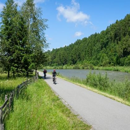 Cyclists on paved path along Dunajec River with wooden fence, green meadows, and forested hills under blue sky.