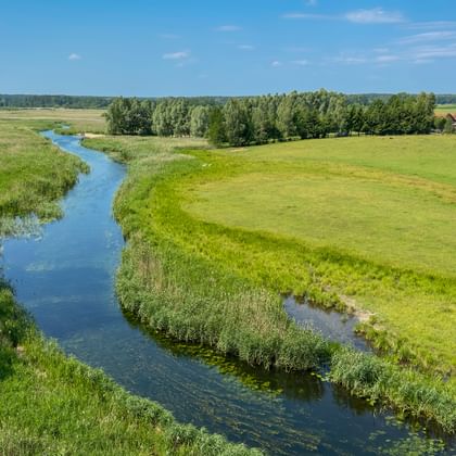 Luftaufnahme eines blauen Flusses, der sich durch grüne Wiesen bei Wojnowo-Kruttina in Masuren schlängelt, mit Bäumen und Bauernhaus.