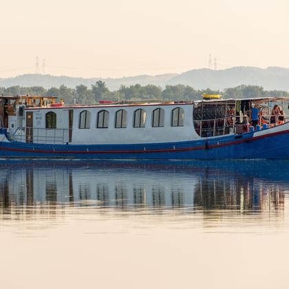 MS Lestello, ein blau-weißes Flusskreuzfahrtschiff mit Bogenfenstern, fährt auf ruhigem Wasser mit sichtbarer Spiegelung. Hügel und Bäume im Hintergrund.