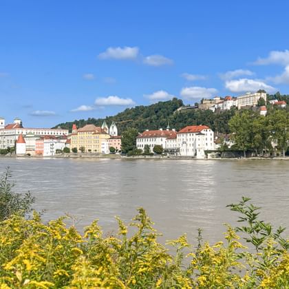 Blick über die Donau auf Passau mit historischen Gebäuden und Kirchen am Ufer, Hügel mit Gebäuden im Hintergrund, blauer Himmel.