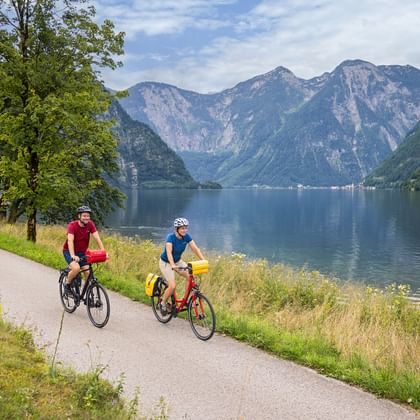 Zwei Radfahrer auf einem Seeweg am Hallstättersee im Salzkammergut, Österreich, mit Bergen im Hintergrund und ruhigem blauen Wasser.