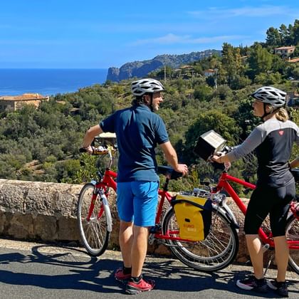 Two cyclists with helmets and red touring bikes with yellow panniers studying a map on a coastal road in Mallorca, overlooking the blue sea.