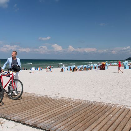 Zwei Radfahrer schieben ihrer Räder auf einem Holzsteg am Strand, im Hintergrund Strandkörbe
