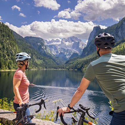 Two cyclists with helmets pause at Gosausee lake shore, viewing the Dachstein mountain range with snow-capped peaks and forested slopes.