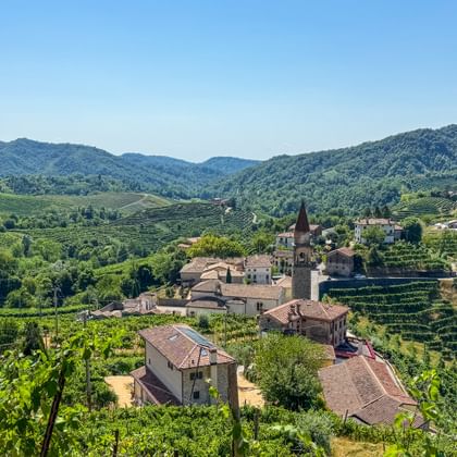 Kleines italienisches Dorf mit Kirchturm, umgeben von terrassierten Prosecco-Weinbergen bei Conegliano, mit sanften Hügeln im Hintergrund.