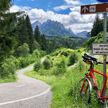 Cycle break with bike leaning against a road sign, with beautiful cycle path in the foreground and the Carnic Alps in the background