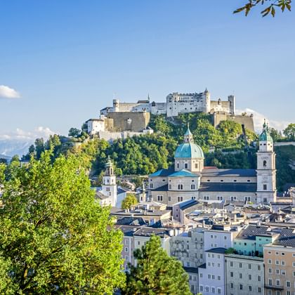 Salzburg cityscape with historic buildings, baroque church domes, and Hohensalzburg Fortress on a hilltop. Green trees frame the view under blue sky.