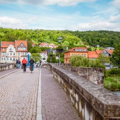 Radfahrer überqueren historische Steinbrücke in Hann. Münden mit bunten Fachwerkhäusern und grünen Hügeln im Hintergrund.