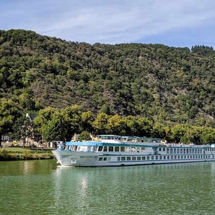 MS Switzerland Flusskreuzfahrtschiff fährt auf ruhigem grünen Wasser mit bewaldeten Hügeln im Hintergrund unter teilweise bewölktem Himmel.