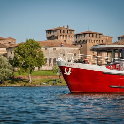 Red and white passenger ship MS Ave Maria on blue water with historic fortress towers and green trees along the waterfront in Mantua.