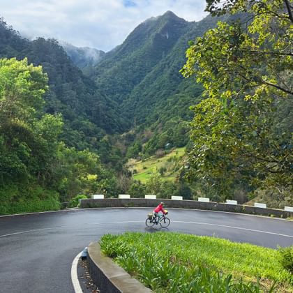 Radfahrer in roter Jacke auf kurvenreicher Bergstraße nahe Gambao, Madeira, mit grünem Tal und dramatischen Berggipfeln im Hintergrund.