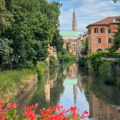 Canal in Vicenza with red flowers in foreground, historic buildings and green copper-roofed church with tall bell tower reflected in water.