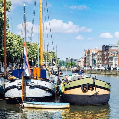 Traditionelle Segelboote im Kanal von Dordrecht mit historischen niederländischen Gebäuden am Ufer unter blauem Himmel.