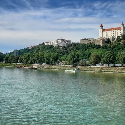 Bratislava Castle with white walls and red roofs on a green hill above the Danube River. Ships are moored along the tree-lined riverbank.