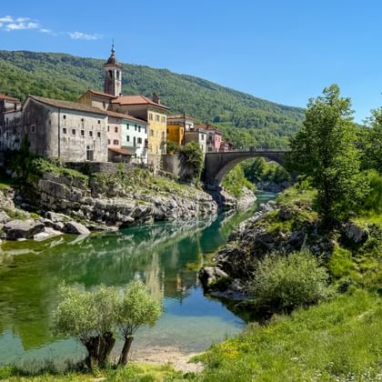Historic stone bridge spanning a green river in Kanal ob Soči, Slovenia, with colorful buildings and forested hills in the background.