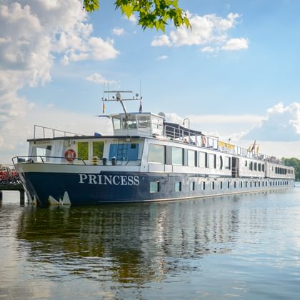 River cruise ship MS Princess moored at a wooden pier on calm water. The white vessel reflects in the water under a blue sky with clouds.