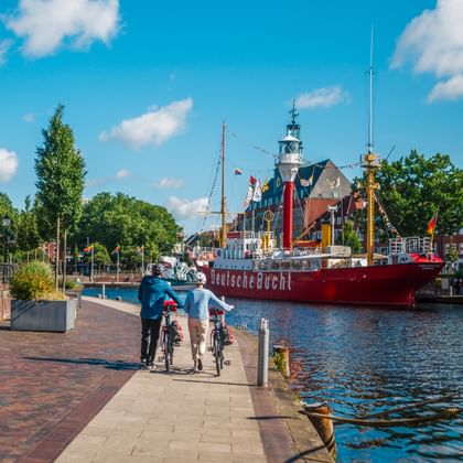 Zwei Radfahrer fahren entlang eines gepflasterten Uferwegs mit rotem Feuerschiff und Leuchtturm im Hafen unter blauem Himmel mit weißen Wolken.