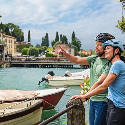 Zwei Radfahrer mit Helmen am Hafen von Peschiera am Gardasee, mit Booten im türkisfarbenen Wasser und italienischen Gebäuden mit Zypressen.