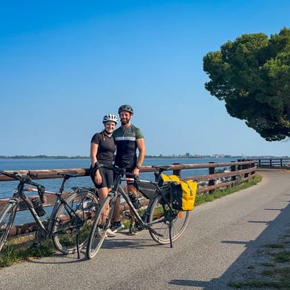 Two cyclists with helmets and touring bikes on coastal path with wooden fence, lagoon, and pine tree near Grado on the Alpe Adria cycle route.