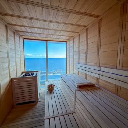 Light wood sauna interior with tiered benches and heater, featuring large window overlooking blue ocean in Porto Moniz, Madeira.