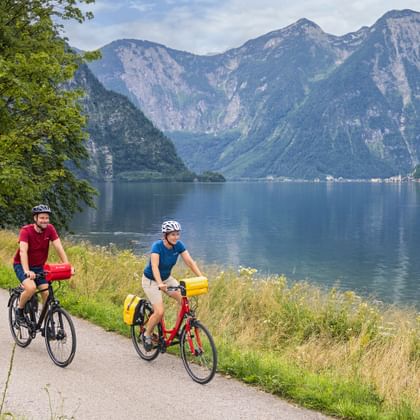 Two cyclists riding on a paved path along Lake Hallstatt with mountains in the background and calm blue water beside the trail.
