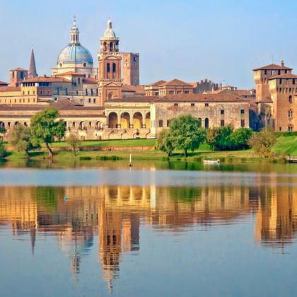 Historische Skyline von Mantua mit Domkuppel und Glockenturm spiegelt sich in ruhigem Wasser. Grüne Bäume säumen das Ufer mit terrakottafarbenen Gebäuden.