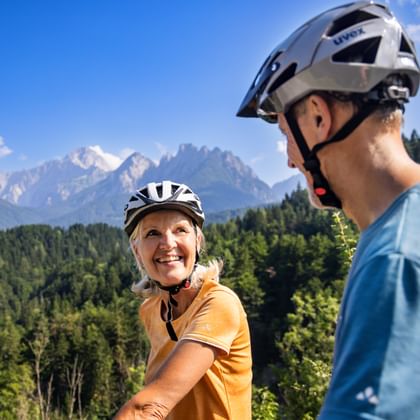 Zwei Radfahrer mit Helmen genießen Bergblick bei Tarvis. Bewaldete Hügel und dramatische Alpengipfel unter blauem Himmel im Hintergrund.