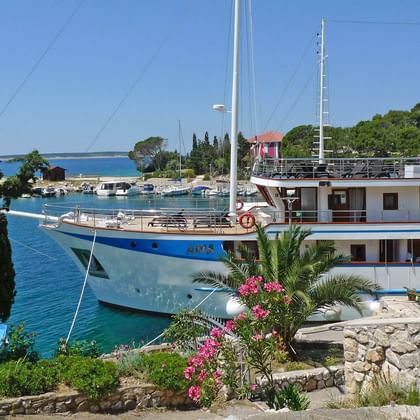 Weiße und blaue Motoryacht MS Aria mit zwei Masten im Mittelmeerhafen. Steinmauern mit rosa Blumen rahmen den Blick auf das türkisfarbene Wasser.