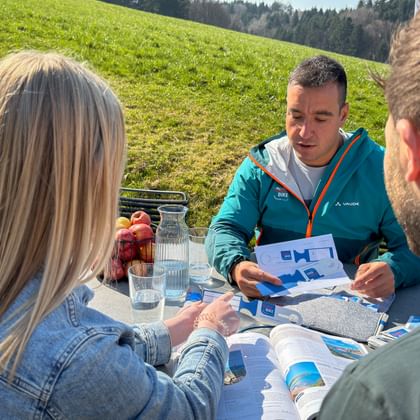 Beratungsgespräch im Freien mit Eurobike-Mitarbeiter in türkisfarbener Jacke, der zwei Gästen Broschüren an einem Tisch auf einer sonnigen Wiese zeigt.