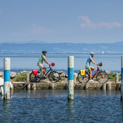 Two cyclists riding along a wooden pier in Grado harbor with boats moored on both sides, mountains visible in the background under blue sky.