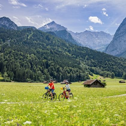 Zwei Radfahrer umgeben von Blumenwiesen und Almhütten, im Hintergrund das Zugspitzmassiv bei Garmisch-Partenkirchen