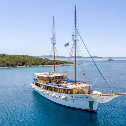 White two-masted sailing vessel MS Macek anchored in calm turquoise waters near a green forested coastline under clear blue sky.